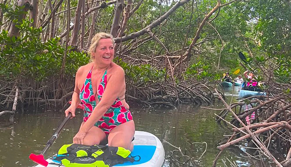 A person is sitting on a paddleboard amidst mangroves looking back with a smile while others kayak behind her