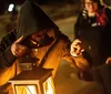 A hooded figure holds a lit lantern pointing forward with a focused look in a dimly lit outdoor setting as another person watches from the background