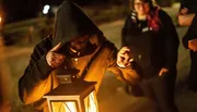 A hooded figure holds a lit lantern, pointing forward with a focused look in a dimly lit outdoor setting, as another person watches from the background.