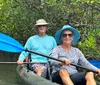 Two people are smiling while kayaking among mangroves both holding blue paddles and wearing sun hats