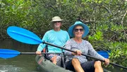 Two people are smiling while kayaking among mangroves, both holding blue paddles and wearing sun hats.