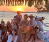 A group of cheerful people is posing for a photo on a sunny day at a waterfront location with a thatched roof structure extending their arms and smiling