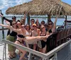 A group of cheerful people is posing for a photo on a sunny day at a waterfront location with a thatched roof structure extending their arms and smiling