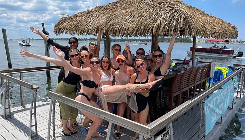 A group of cheerful people is posing for a photo on a sunny day at a waterfront location with a thatched roof structure extending their arms and smiling