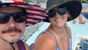 Two people are smiling for a selfie on a boat, wearing sunglasses and wide-brimmed hats.