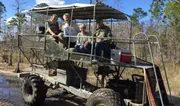 Four individuals are sitting on a large, open-air swamp buggy vehicle in a wooded area.