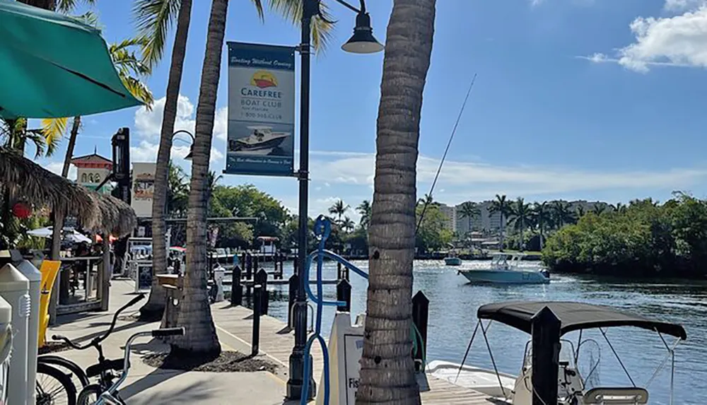 A sunny waterfront scene with palm trees and a boat passing by adjacent to a dock lined with amenities and a sign for a boat club