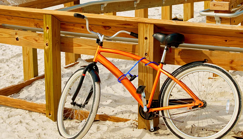 An orange bicycle is casually leaned against a wooden railing on a sandy beach under bright sunlight