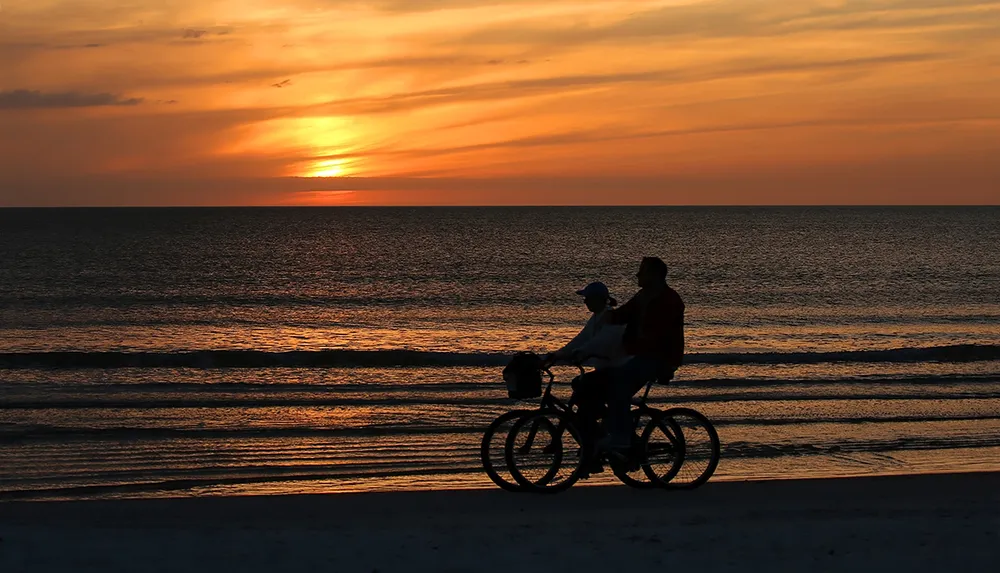 Two people are riding a tandem bicycle along the beach at sunset creating silhouettes against the vibrant sky