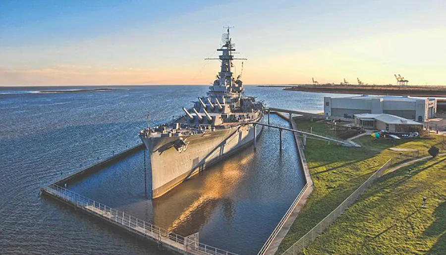 A retired battleship is docked as a museum next to a calm body of water under a clear sky at sunset.