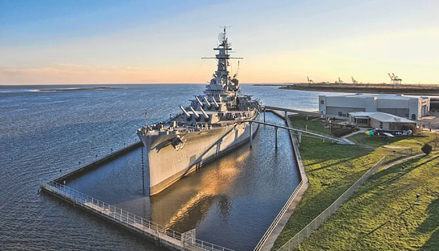 The image shows a large battleship moored in a water dock against a backdrop of open water and a clear sky during sunset or sunrise.