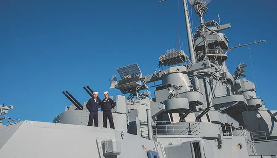 Two individuals are standing in naval attire on the deck of a naval ship with large gun turrets and radar equipment against a clear blue sky.