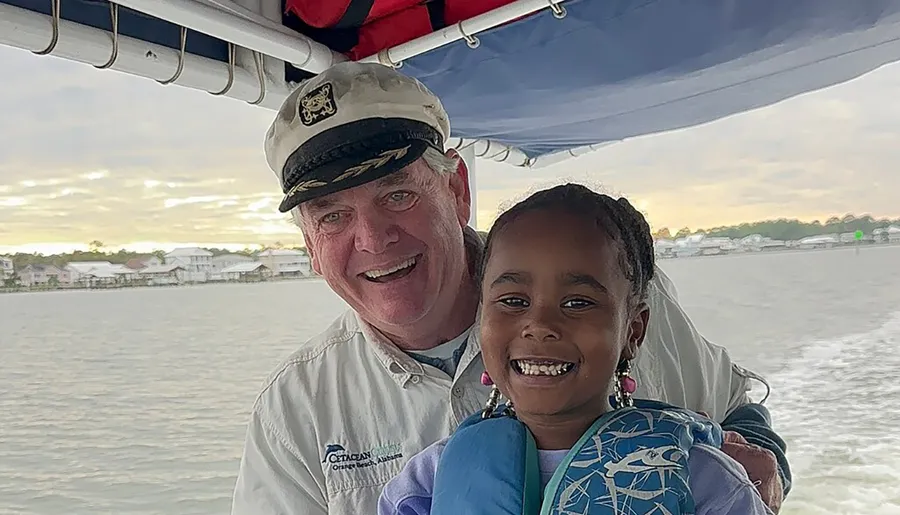 An adult in a captain's hat and a smiling child wearing a life jacket pose for a photo on a boat with waterfront houses in the background.