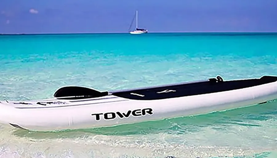 A paddleboard labeled TOWER rests on the clear, turquoise waters of a tranquil beach with a sailboat in the distance.