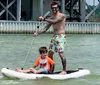 A tattooed adult is standing and paddling on a paddleboard while a child sits in front both appearing to enjoy a day on the water