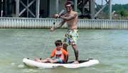 A tattooed adult is standing and paddling on a paddleboard while a child sits in front, both appearing to enjoy a day on the water.