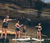 Four individuals are standing on paddleboards engaging in an activity near a rocky cliff with a small waterfall