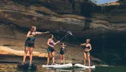 Four individuals are standing on paddleboards, engaging in an activity near a rocky cliff with a small waterfall.