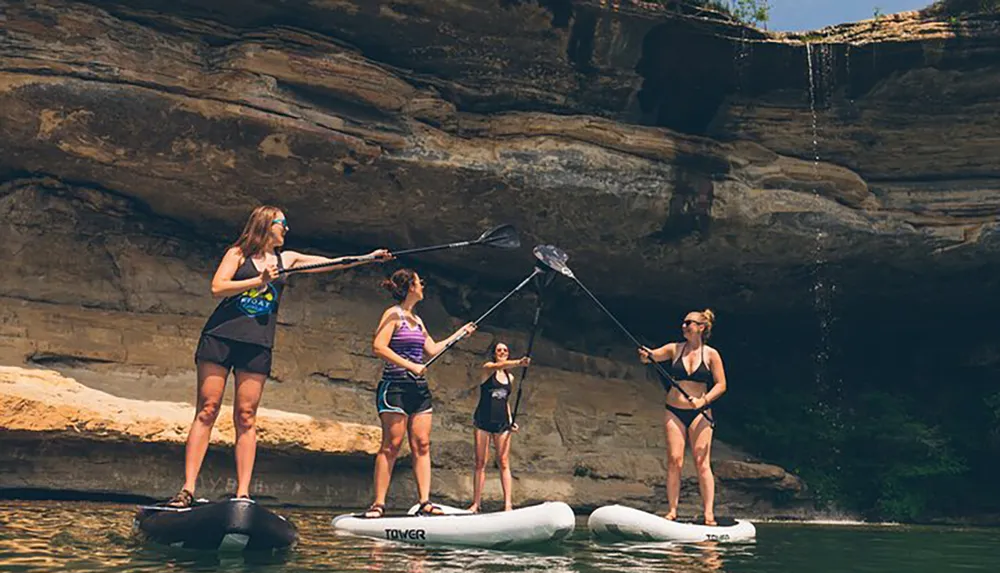 Four individuals are standing on paddleboards engaging in an activity near a rocky cliff with a small waterfall