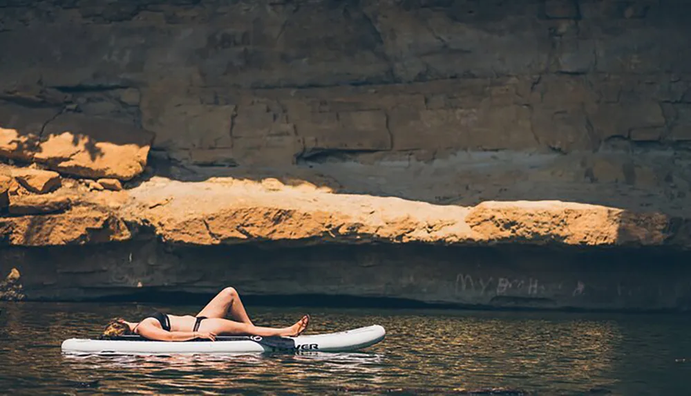 A person is relaxing on an inflatable raft in calm waters near a rocky cliff