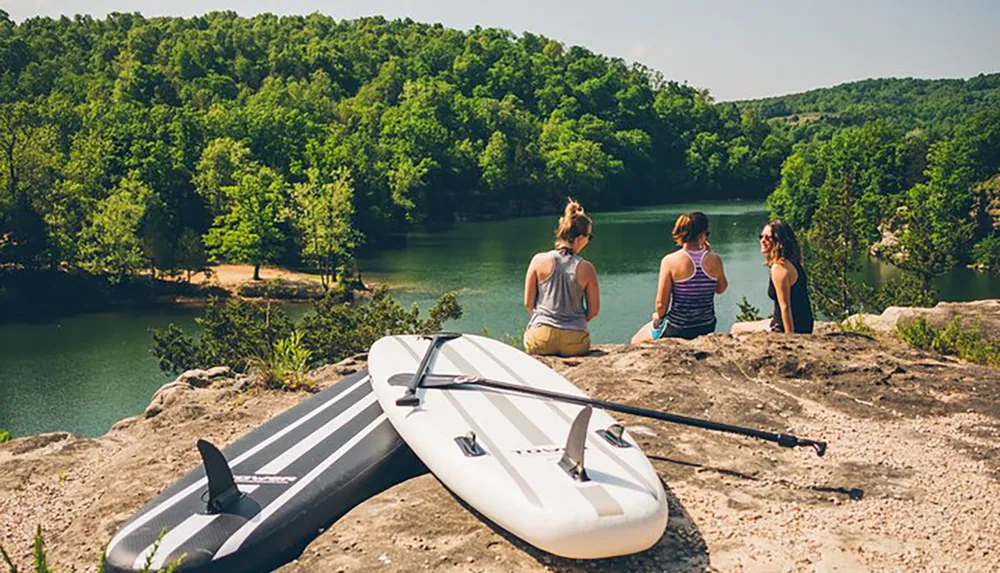 Three people are sitting on a rocky outcrop overlooking a serene lake with a paddleboard beside them