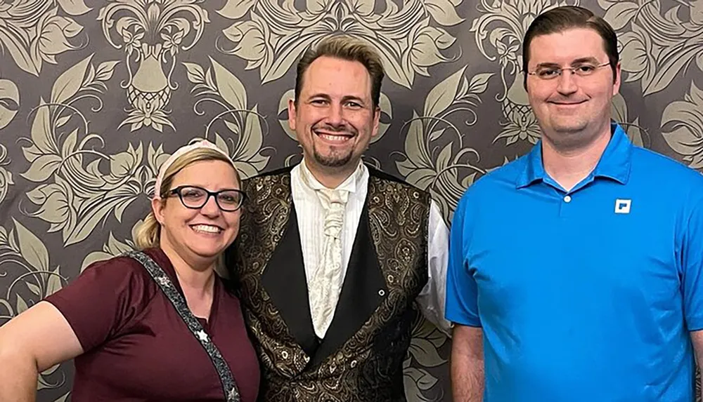 Three people are smiling for the camera with the person in the middle wearing a more formal attire with a patterned waistcoat flanked by a woman to the left and a man to the right against a wallpapered backdrop