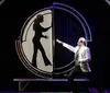 A man in a suit gestures towards a large circular screen displaying the silhouette of a woman in a pose in what appears to be a theatrical or magic show setting