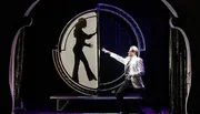 A man in a suit gestures towards a large circular screen displaying the silhouette of a woman in a pose, in what appears to be a theatrical or magic show setting.