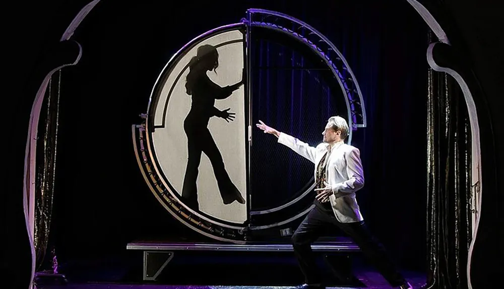 A man in a suit gestures towards a large circular screen displaying the silhouette of a woman in a pose in what appears to be a theatrical or magic show setting