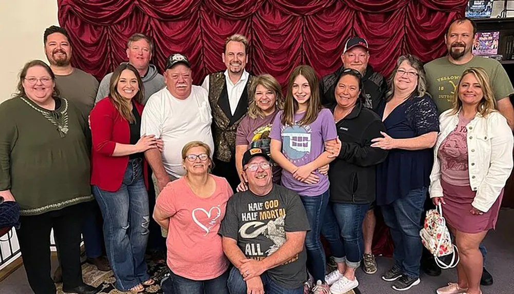 The image shows a group of smiling people posing together for a photo in front of a red draped background appearing to be gathered for a social or family event