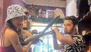 Two people are playfully posing with oversized props as swords inside a room filled with various colorful decorations and novelty items.