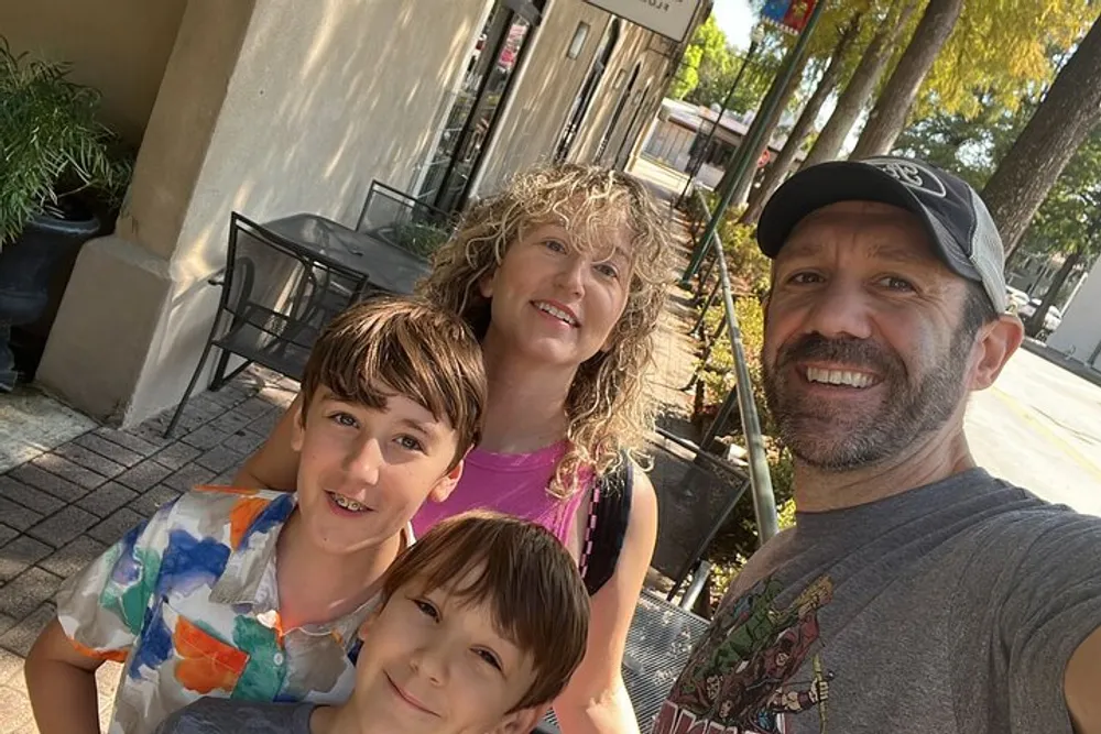 A family of four is smiling for a selfie on a sunny day on a city sidewalk