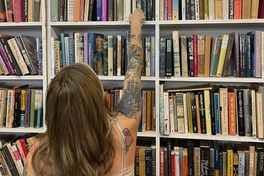 A person with tattooed arms is reaching out for a book on a shelf filled with an array of books