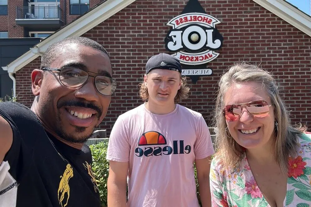 Three people are taking a group selfie outside in front of a building with a sign reading HOTEL in reversed letters