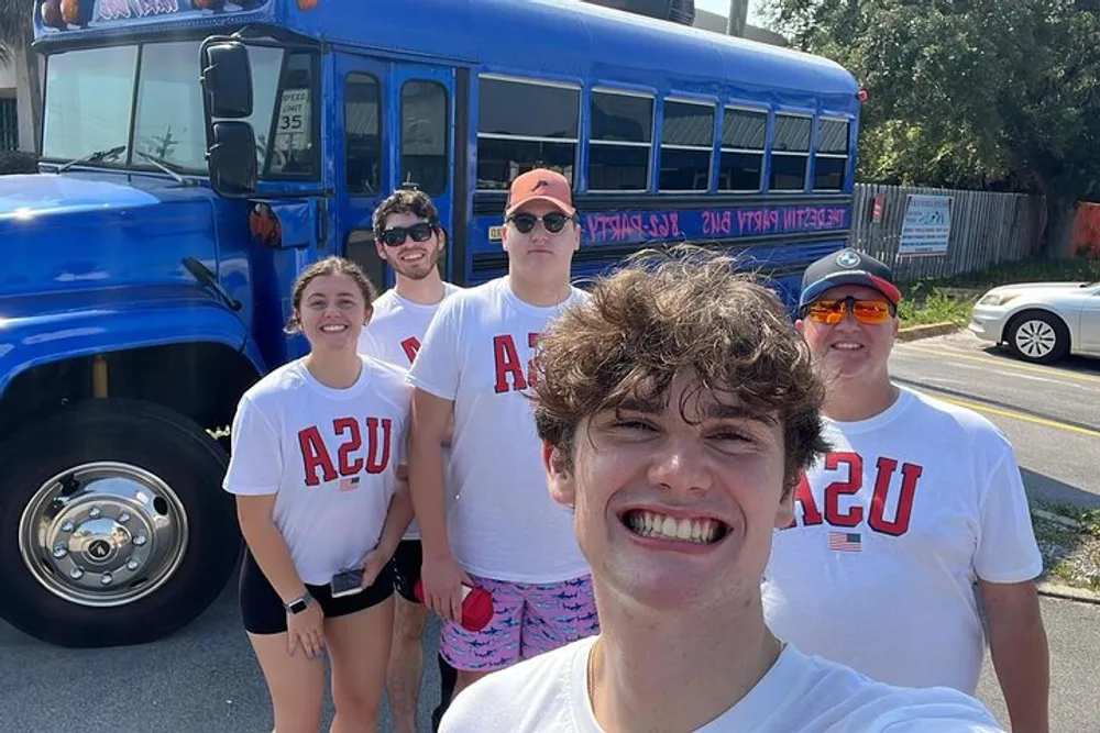 A group of five individuals is posing for a selfie in front of a blue bus all wearing matching white t-shirts with red AZU lettering appearing to be in a cheerful mood