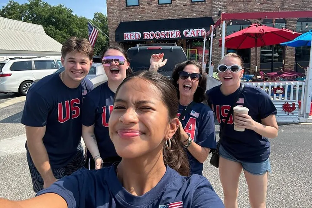 A group of cheerful people wearing USA shirts are taking a selfie outside the Red Rooster Cafe under a sunny sky