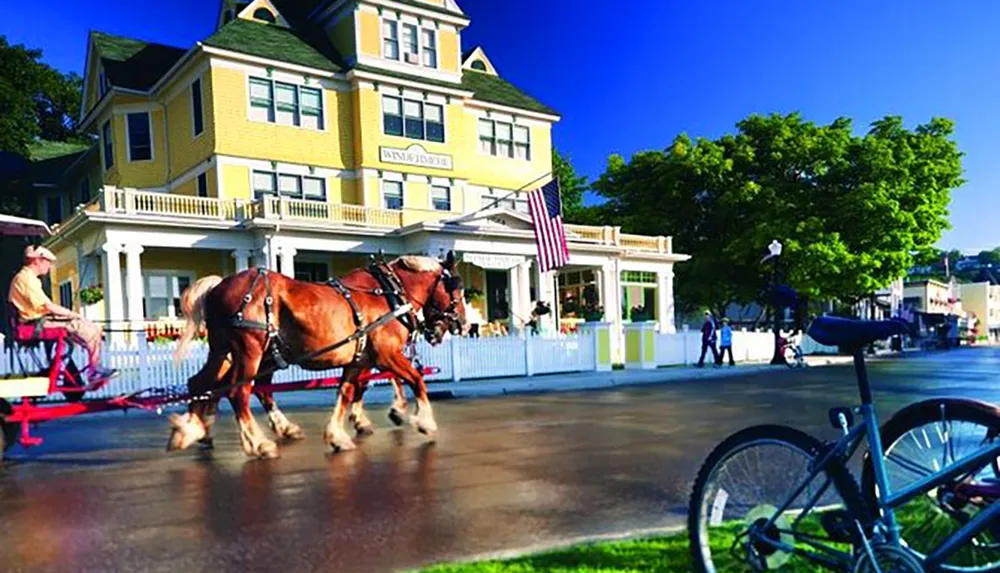 A horse-drawn carriage travels past a yellow building with an American flag on a sunny day while bicycles are parked nearby