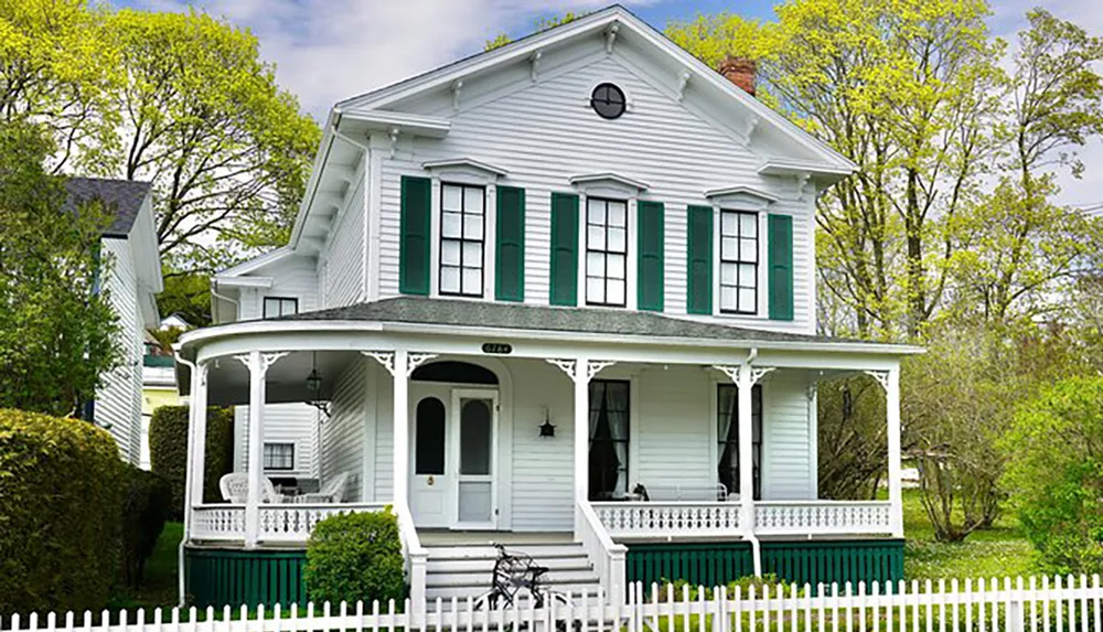 The image showcases a charming two-story white house with green shutters and a wrap-around porch enclosed by a white picket fence conveying a classic American home aesthetic