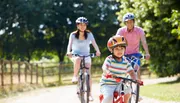 A child leads the way on a bike ride with two adults cycling behind on a sunny, tree-lined path.