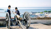 Two individuals dressed in historic military-style uniforms are standing near an old cannon overlooking a scenic coastal town and harbor.