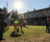 The image shows an outdoor community event with individuals engaging in juggling and playing the violin on a grassy lawn with a historic building and spectators in the background bathed in the warm glow of a setting sun