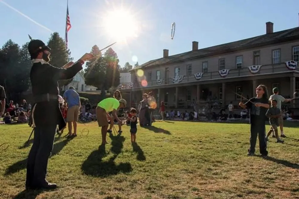 The image shows an outdoor community event with individuals engaging in juggling and playing the violin on a grassy lawn with a historic building and spectators in the background bathed in the warm glow of a setting sun