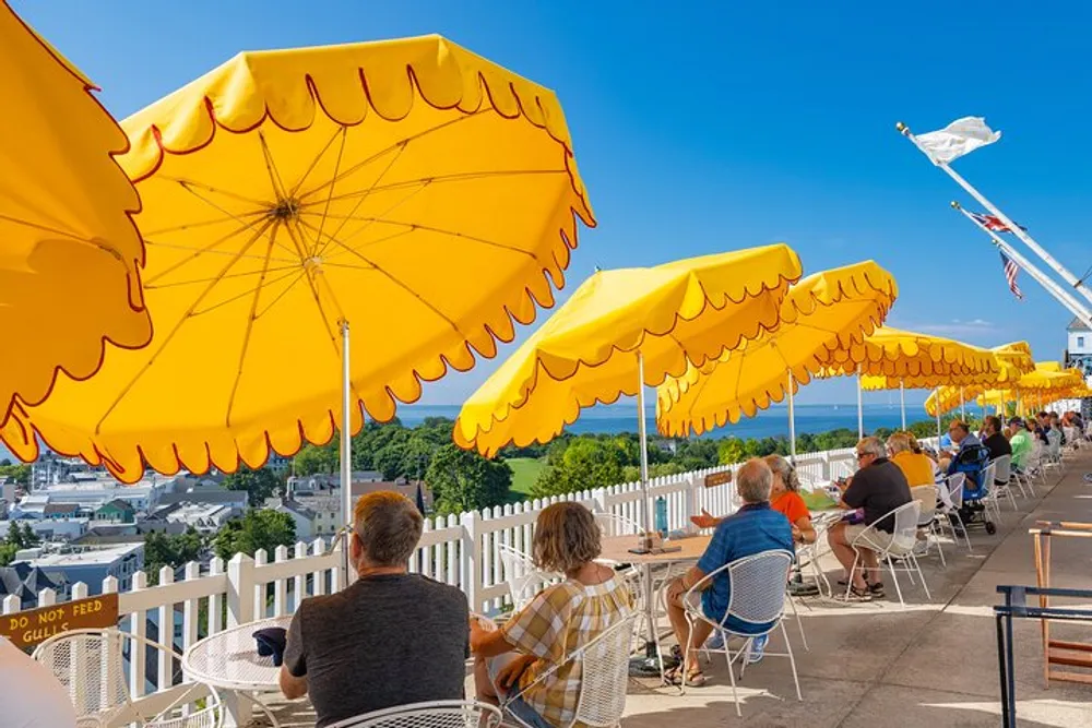 People are seated at an outdoor patio under bright yellow umbrellas enjoying a sunny day with a view of a picturesque landscape and a flag flying in the background