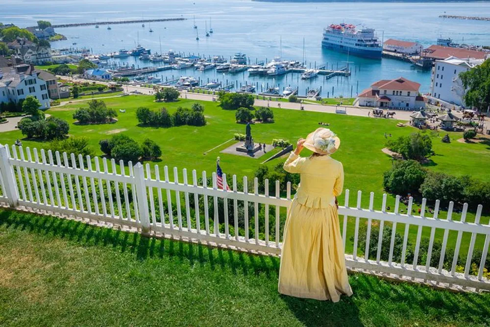 A person in historical clothing overlooks a picturesque coastal town with a marina and green parklands from behind a white picket fence