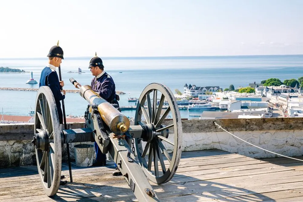 Two individuals in ceremonial military attire are conversing beside an old cannon with a panoramic view of a coastal town and body of water in the background