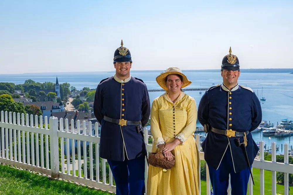 Two individuals in historical military uniforms flank a person in a yellow period dress standing in front of a scenic overlook with a view of the water and a town below