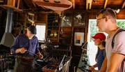 A blacksmith is working on a piece of metal with a hammer as two onlookers watch attentively in a rustic workshop setting.