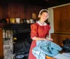 A woman in historical dress is smiling at the camera while standing in a rustic kitchen with a fireplace and antique cookware