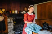 A woman in historical dress is smiling at the camera while standing in a rustic kitchen with a fireplace and antique cookware.