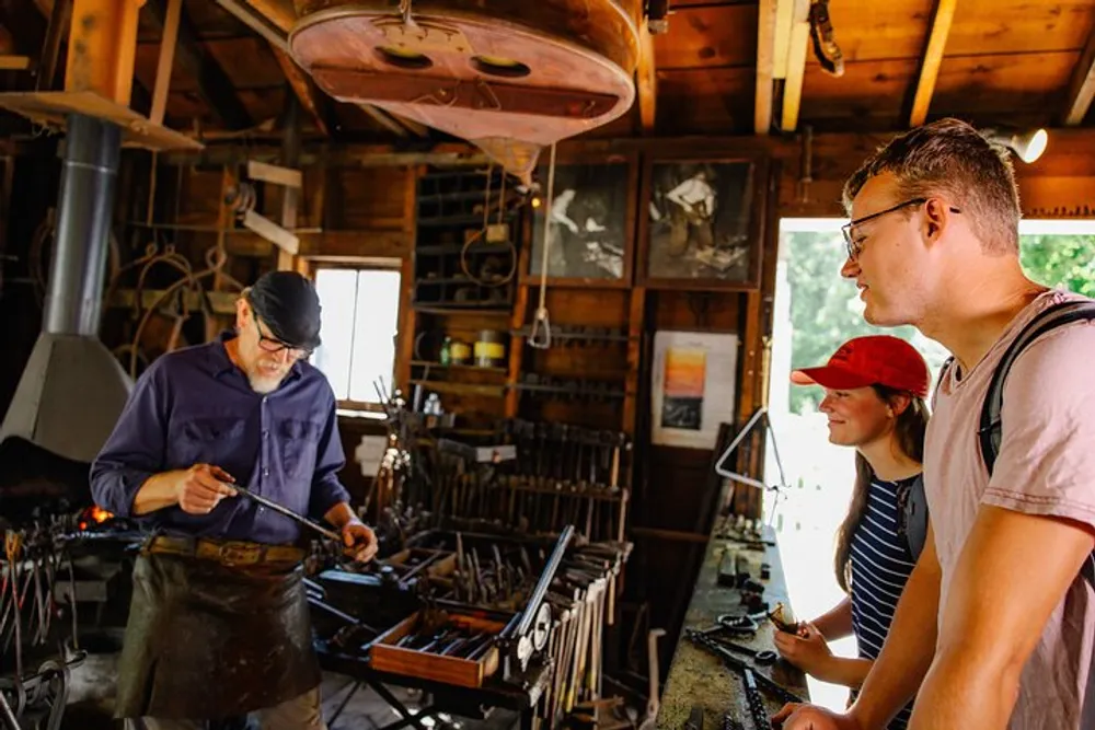 A blacksmith is working on a metal piece at a forge while two onlookers watch with interest